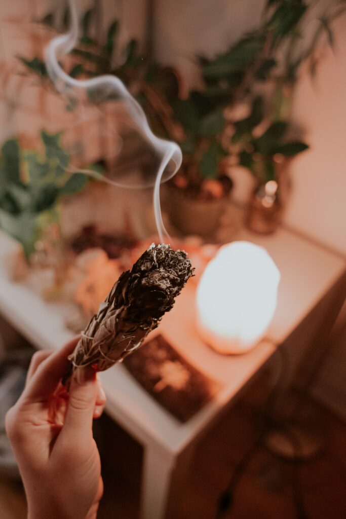 Close-up of a hand holding a smoldering sage bundle with curling smoke indoors, plants blurred in the background, warm lighting