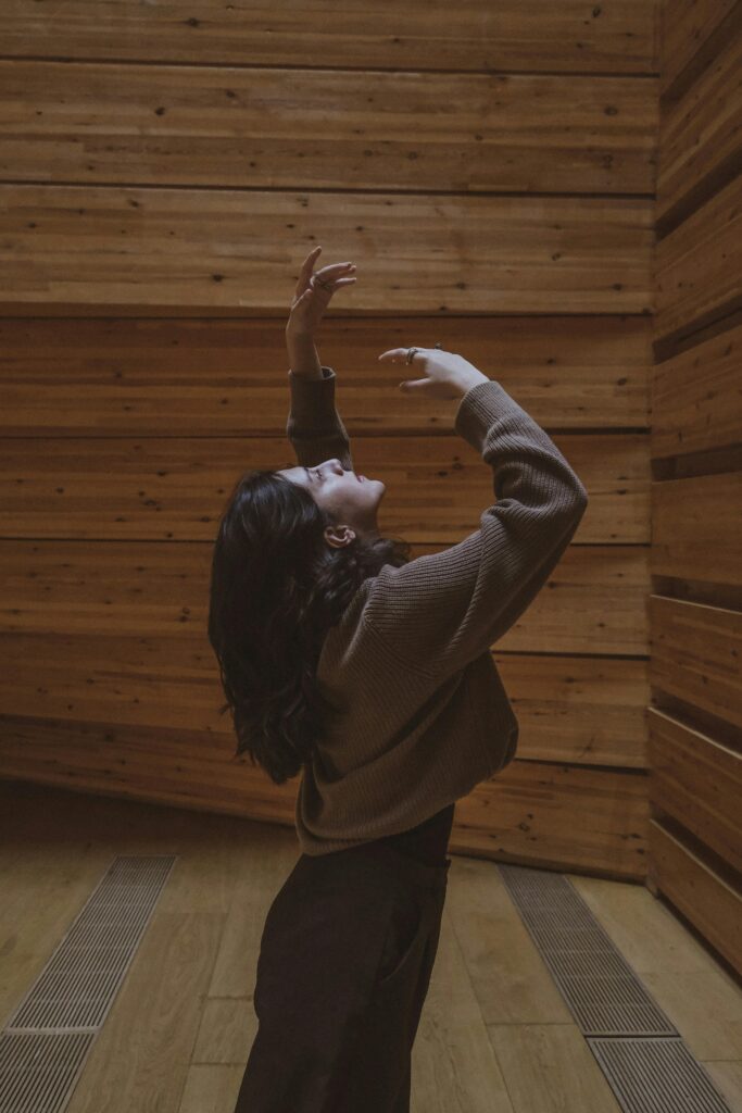 pexels-filiamariss-14043017 | SoulCare Studios Person with dark hair wearing a brown knit sweater raises arms in a wooden-paneled room, mid-dance or pose.